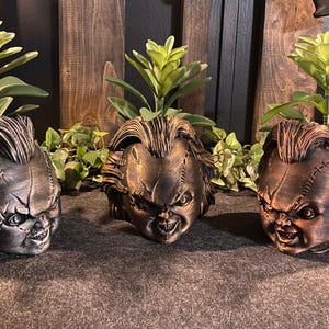 May include: Three sculpted planters shaped like the head of a doll with stitched facial features and messy hair. Each planter holds a green succulent plant. The planters are finished in metallic silver, bronze, and copper tones respectively.