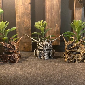 May include: Three decorative planters shaped like robot heads, each with a different colour scheme: gold, silver and black. Each planter has a small succulent plant growing inside.