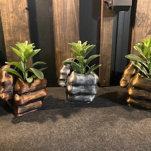 May include: Three potted plants in unique, hand-shaped planters. The planters are made of a textured material and are painted in gold, silver, and bronze.