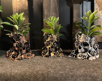 May include: Three decorative skull planters with faux succulents. The planters are intricately detailed with rose designs and painted in shades of bronze, gold, and silver. The plants have green leaves.