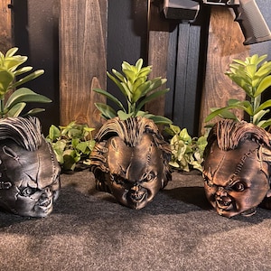 May include: Three ceramic planters shaped like the head of a fictional horror character with stitched-up skin and a menacing expression. Each planter is decorated with a different color scheme, including black, gold, and brown. The planters are filled with small green plants.