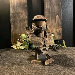 May include: A bronze-effect bust of a futuristic soldier, with a helmet and armour. The sculpture is mounted on a hexagonal base. The bust has detailed, textured surfaces and is set against a dark wooden background.