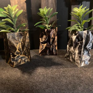 Puede incluir: Tres maceteros decorativos con vegetación artificial. Los maceteros están hechos de papel arrugado texturizado en tonos marrones y grises. Las plantas tienen hojas verdes. Los maceteros están colocados sobre una superficie gris oscuro.