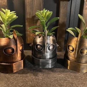 May include: Three robot head planters in copper, silver, and gold metallic finishes, each containing a green succulent plant. The planters feature a central vertical ridge, horizontal eye slits, and a rounded base with side bolt details.