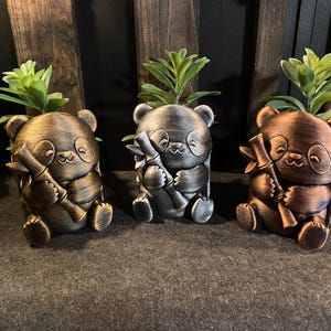 May include: Three panda-shaped planters in metallic bronze, silver, and copper finishes. Each panda figure is seated, holding a stalk of bamboo, and contains a small green succulent plant. The figures feature rounded ears, closed eyes, and a small smile.