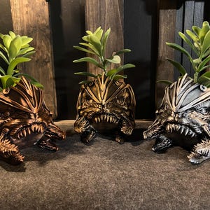 May include: Three ceramic planters shaped like a monster's face, each with a different color finish: gold, bronze, and silver. Each planter has a succulent plant growing inside.