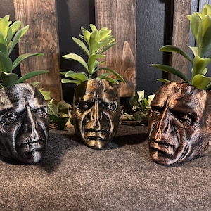 May include: Three 3D printed planters shaped like human heads, each with a different color finish: black, bronze, and gold. Each planter has a small succulent plant growing inside.