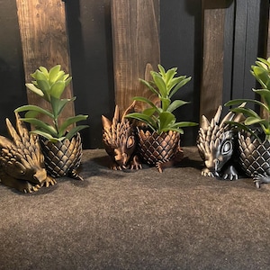 Könnte beinhalten: Drachenförmige Pflanzgefäße in Bronze- und Silbertönen, die jeweils eine kleine grüne Pflanze enthalten. Die Pflanzgefäße sind mit Schuppen und Drachenmerkmalen detailliert, was ein Fantasy-Thema schafft.