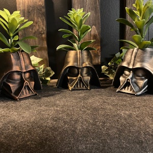 Puede incluir: Tres macetas de Darth Vader de bronce oscuro con suculentas verdes que crecen en su interior. Las macetas están hechas de un material liso de bronce oscuro y tienen un diseño detallado de Darth Vader.