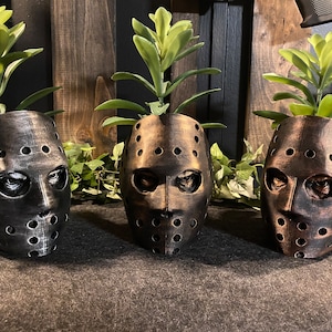 Può includere: Tre vasi a forma di maschera da hockey nera e bronzo con piante verdi che crescono al loro interno. Le maschere hanno un aspetto usurato e sono un accessorio popolare per i film horror.