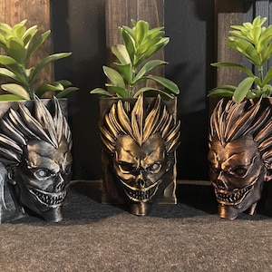 May include: Three decorative planters, each featuring a detailed, stylized face design. The planters are in shades of silver, gold, and bronze, and each holds a green plant. The planters are square and have a textured finish.