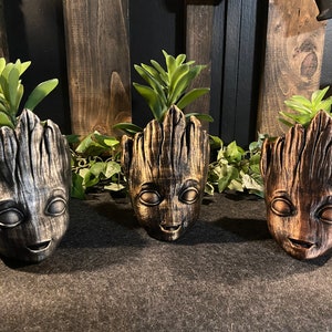 May include: Three potted plants in Groot-shaped planters. The planters are made of a dark gray, gold, and brown material. The plants are green and leafy.