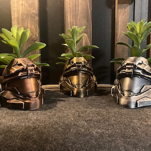 Puede incluir: Tres macetas impresas en 3D con forma de casco de soldado. Los cascos están pintados en un acabado bronce, dorado y plateado. Cada casco tiene una pequeña planta verde que crece en la parte superior.