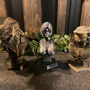 May include: Three detailed sculptures are displayed. The first is a gold and black creature mask. The second is a silver bust of a woman. The third is a gold and black helmet with the number 117. All are on black bases.