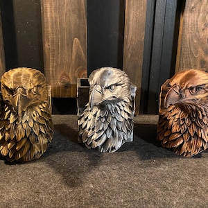 May include: Three decorative eagle head planters in varying colors: gold, silver, and brown. Each planter features a detailed carving of an eagle's head with a stern expression and a textured feather design. The planters are set against a dark wooden background.