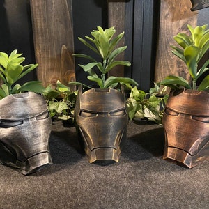 May include: Three Iron Man helmet-shaped planters in metallic finishes: silver, gold, and bronze. Each planter holds a green succulent. The planters are arranged on a dark surface with a wooden background, showcasing a unique home decor item.