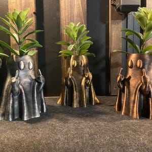 May include: Three ghost-shaped planters in different metallic finishes: silver, gold, and bronze. Each planter has two eyes and is giving the middle finger. Each planter has a small succulent plant growing inside.