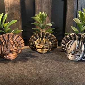 Puede incluir: Tres macetas decorativas con forma de cara de personaje de dibujos animados con un sombrero de paja. Las macetas son de metal y están pintadas en oro, bronce y plata. Cada maceta tiene una pequeña planta creciendo en su interior.