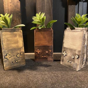 May include: Three wooden planters shaped like vintage handheld game consoles. Each planter is a different color: brown, gold, and gray. The planters are filled with small green plants.