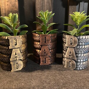 May include: Three decorative planters made from repurposed tires, each holding a succulent plant. The planters are painted in shades of brown, bronze, and blue, with the word "DAD" attached to the front of each planter.