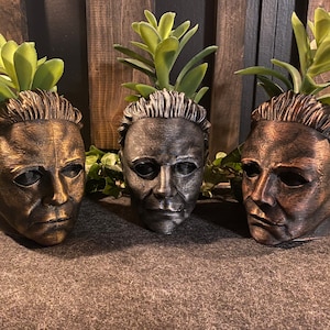 May include: Three Halloween-themed planters, each featuring a different coloured Michael Myers mask. The masks are painted in gold, silver, and copper. Each planter has a small succulent plant growing out of the top.