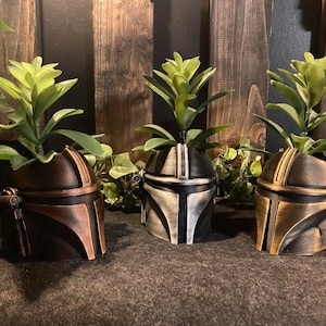 May include: Three potted plants in different colored Mandalorian helmets. The helmets are made of plastic and have a weathered look. The plants are green and leafy.