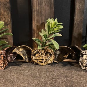 May include: Three ceramic planters shaped like the character Gizmo from the movie Gremlins. Each planter is a different color: gold, bronze, and silver. Each planter has a small green plant growing out of the top.