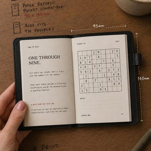 May include: A black leather-bound notebook open to a Sudoku puzzle page. The left page displays instructions titled How to Play, while the right page shows a grid labeled Puzzle 01. The notebook measures 95 mm by 140 mm, as indicated by text.