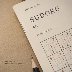 May include: A beige Sudoku puzzle booklet titled EASY COLLECTION 001 with 25 EASY PUZZLES printed on the cover. The booklet features a partial Sudoku grid with red numbers and a black pencil tip visible at the bottom edge of the paper cover.