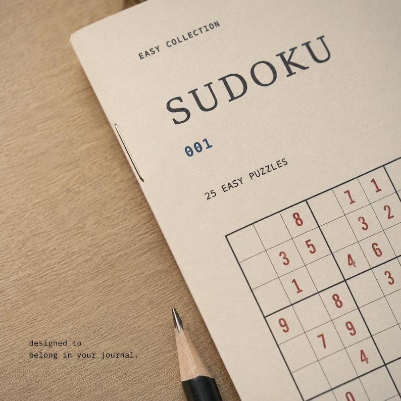 May include: A beige Sudoku puzzle booklet titled EASY COLLECTION 001 with 25 EASY PUZZLES printed on the cover. The booklet features a partial Sudoku grid with red numbers and a black pencil tip visible at the bottom edge of the paper cover.