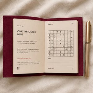 May include: An open Sudoku puzzle book with a deep red fabric cover and a gold pen. The left page displays instructions titled How to Play, while the right page shows a grid labeled Puzzle 01 Easy. The text includes One Through Nine and A Note From The Test Lab.