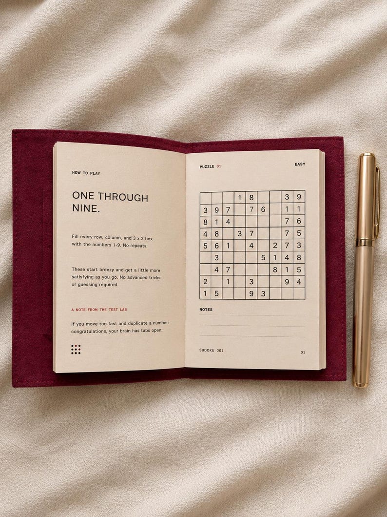 May include: An open Sudoku puzzle book with a deep red fabric cover and a gold pen. The left page displays instructions titled How to Play, while the right page shows a grid labeled Puzzle 01 Easy. The text includes One Through Nine and A Note From The Test Lab.