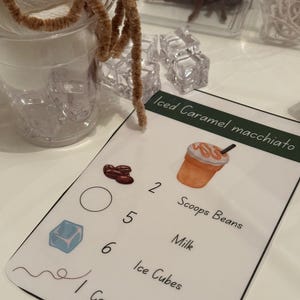 Puede incluir: Una tarjeta de receta imprimible para Iced Caramel Macchiato con una ilustración de dibujos animados de la bebida. La tarjeta enumera los ingredientes y las cantidades necesarias: 2 cucharadas de granos de café, 140 ml de leche, 6 cubitos de hielo y 1 remolino de caramelo.