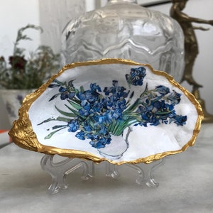 May include: A white oyster shell with a gold rim, painted with a bouquet of blue flowers in a vase. The shell is on a clear plastic stand.