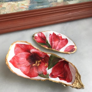 Op de afbeelding: Twee oesterschelpen met gouden randen, beschilderd met rode amaryllis bloemen. De schelpen zijn een decoratief accessoire voor thuis.