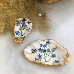 以下が含まれることがあります： 金色の縁取りが施された装飾的なカキの貝殻の皿が2つ。1つの皿には、青い花とハチが描かれた花柄のデザインが施されています。もう1つの皿には、青い花が描かれた花柄のデザインが施されています。