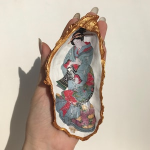 以下が含まれることがあります： 金色の塗料で塗られた牡蠣の殻に、青と赤の着物姿の女性が印刷されています。女性は扇子を持っており、穏やかな表情をしています。