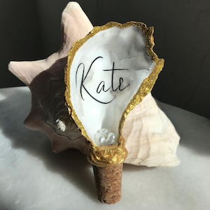 May include: Gold-painted oyster shell bottle stopper with the name "Kate" written inside. The shell is filled with small white pearls and sits on a cork base.