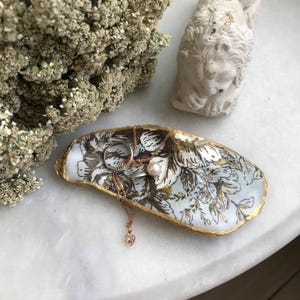 May include: A white oyster shell dish with gold accents and a floral pattern. The dish is filled with jewelry, including a pearl necklace and a ring.