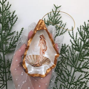 Può includere: Un ornamento a forma di conchiglia di ostrica dipinta d'oro con un dipinto della Nascita di Venere all'interno. L'ornamento ha una corda dorata per appenderlo.