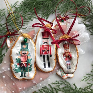 May include: Three handmade Christmas decorations shaped like oyster shells with a gold trim. Each decoration features a painted illustration of a Nutcracker soldier in different poses. The decorations are adorned with red and green ribbons and bells.