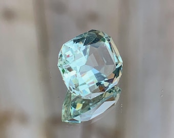 May include: A light blue gemstone with a square cut and many facets. The gemstone is sitting on a reflective surface.