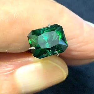 Puede incluir: Una piedra preciosa rectangular facetada de color verde bosque profundo, montada en un engaste de garras de metal plateado. La piedra presenta bordes afilados y limpios, con una superficie transparente y altamente reflectante.