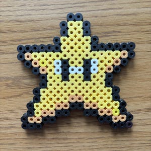 May include: A yellow star-shaped bead art piece with black outlines and white eyes. The star is made of small, colorful beads, and the design is reminiscent of a video game character. The star is placed on a wooden surface.