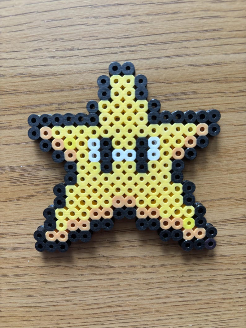 May include: A yellow star-shaped bead art piece with black outlines and white eyes. The star is made of small, colorful beads, and the design is reminiscent of a video game character. The star is placed on a wooden surface.