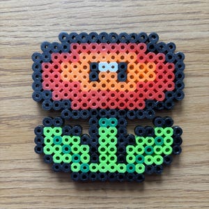 May include: A pixel art flower made from colorful plastic beads. The flower has a red and orange head with black outlines, green stem and leaves, and white eyes. The craft is on a wooden surface.