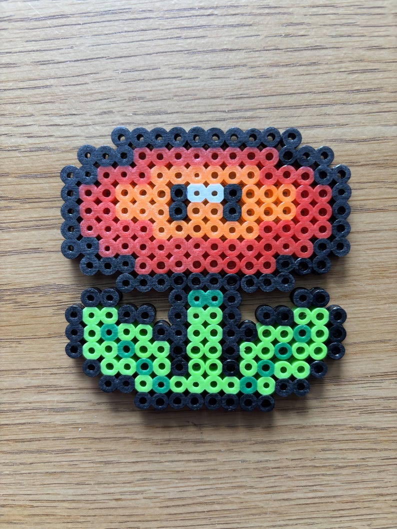 May include: A pixel art flower made from colorful plastic beads. The flower has a red and orange head with black outlines, green stem and leaves, and white eyes. The craft is on a wooden surface.