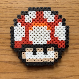 May include: A pixelated mushroom design, crafted from colorful beads. The mushroom features a red cap with white spots, an orange center, and a black base. The design is reminiscent of a classic video game character.