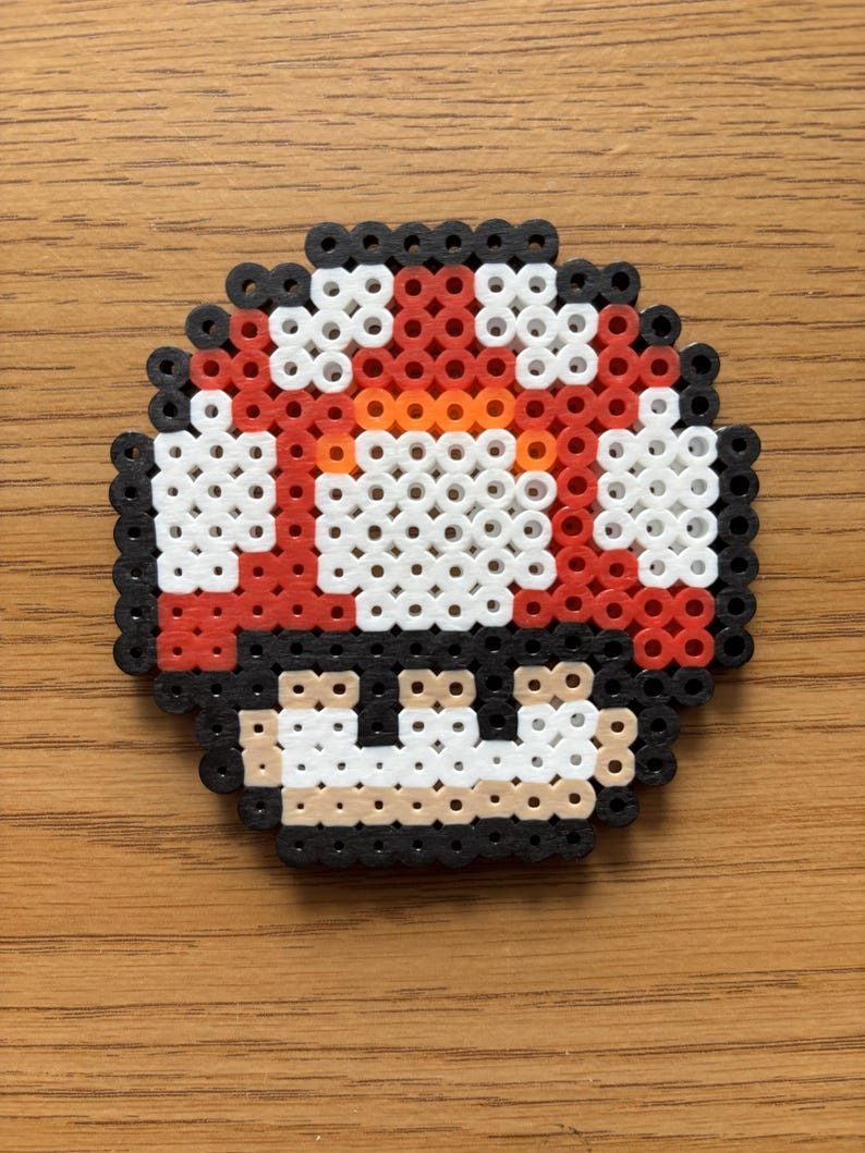 May include: A pixelated mushroom design, crafted from colorful beads. The mushroom features a red cap with white spots, an orange center, and a black base. The design is reminiscent of a classic video game character.
