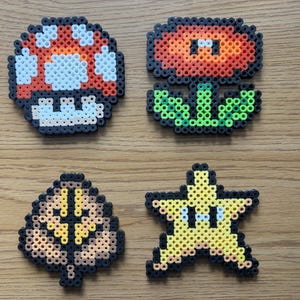 May include: Four pixel art designs from the Super Mario video game series. The designs include a red and white mushroom, a red and orange flower, a brown and yellow leaf, and a yellow star with eyes.